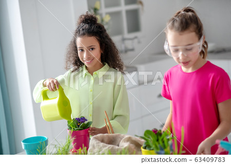 Dark-skinned girl with a watering can, a fair-skinned girlfriend are nearby. 63409783
