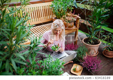 Senior woman with laptop sitting outdoors on terrace, working and eating. Senior woman with laptop sitting outdoors on terrace, working and eating. 63418189