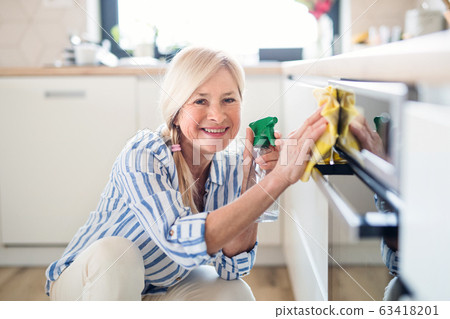 Portrait of senior woman cleaning kitchen cabinet doors indoors at home. Portrait of senior woman cleaning kitchen cabinet doors indoors at home. 63418201