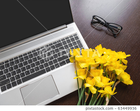 Bouquet of Narcissus or daffodils lying on silver metal laptop. Bright yellow flowers on portable device. Wooden background with glasses. 63449936