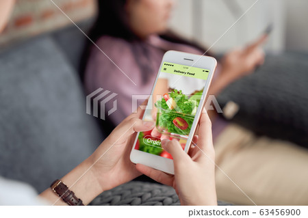Woman using smartphone and touching application screen for ordering salad online at sofa in the home 63456900