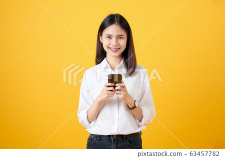 Cheerful beautiful Asian woman holding smartphone and typing message on light yellow background. 63457782