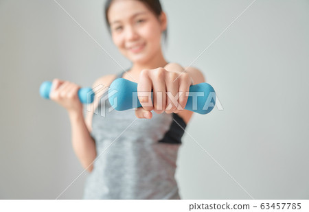 Smiley Asian woman wearing sportswear pumping up muscles with blue dumbbell in living room. 63457785