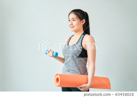 Smiley Asian woman wearing sportswear holding orange mat with blue dumbbell in living room. Smiley Asian woman wearing sportswear holding orange mat with blue dumbbell in living room. 63458028
