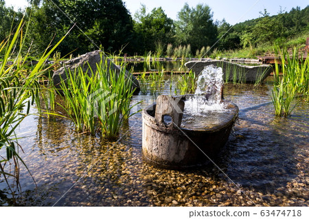 Wooden bucket with oxygenating flowing water at natural swimming pond or pool purifying water without chemicals through biological filters and plants Wooden bucket with oxygenating flowing water at natural swimming pond or pool purifying water without chemicals through biological filters and plants 63474718