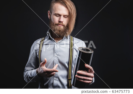 Studio portrait of a bearded stylish long-haired man in a shirt with suspenders. Holding car parts Studio portrait of a bearded stylish long-haired man in a shirt with suspenders. Holding car parts 63475773