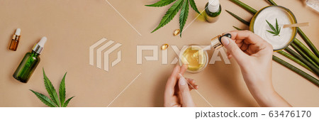 Pipette with CBD cosmetic oil in female hands on a table background with cosmetics, cream with cannabis and hemp leaves, marijuana 63476170