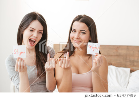 Lesbian couple in bedroom at home sitting holding paper with stigma word grimace cheerful 63477499