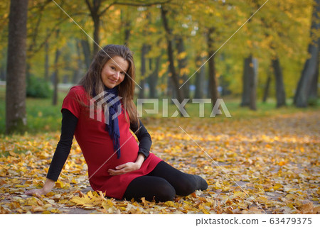 Young attractive pregnant woman in red dress sitting on fallen leaves in autumn park, holding belly. 9 months pregnancy Young attractive pregnant woman in red dress sitting on fallen leaves in autumn park, holding belly. 9 months pregnancy 63479375