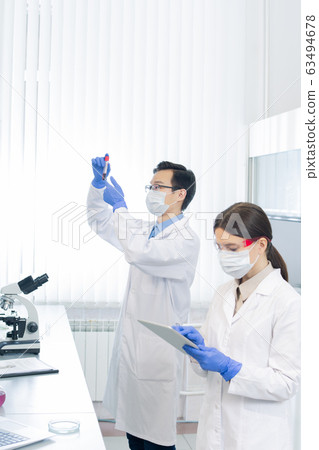 Young female lab worker scrolling in tablet while her colleague looking at flask Young female lab worker scrolling in tablet while her colleague looking at flask 63494678