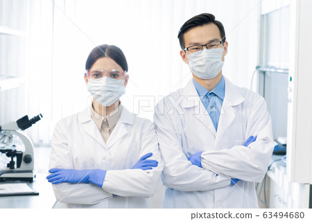 Two young masked intercultural scientists in whitecoats and gloves Two young masked intercultural scientists in whitecoats and gloves 63494680