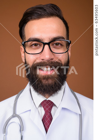 Young handsome bearded Persian man doctor against brown backgrou Young handsome bearded Persian man doctor against brown backgrou 63495603