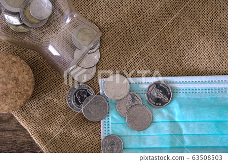 Thai Baht coins poured from glass bottle on gunny sack with blue hygienic mask on wooden table, top view, medical and healthcare fund concept 63508503
