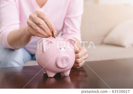 Senior woman putting coin into piggy bank at home Senior woman putting coin into piggy bank at home 63530027