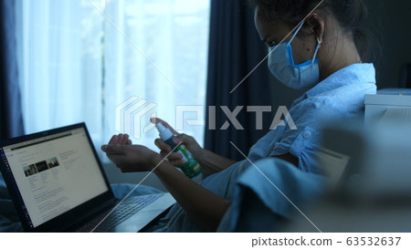 Woman in mask by computer quarantined at home during a pandemic. A woman treats hands with an antiseptic and works on notebook. Woman in mask by computer quarantined at home during a pandemic. A woman treats hands with an antiseptic and works on notebook. 63532637