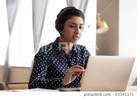 Serious Indian woman wearing headphones talking, using laptop 63545171