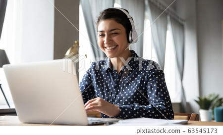 Smiling Indian girl wearing headphones using laptop, working online Smiling Indian girl wearing headphones using laptop, working online 63545173