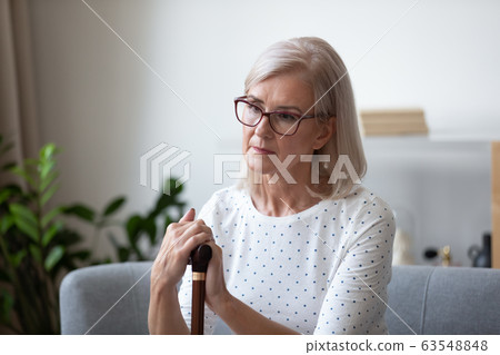Pensive older lady in eyeglasses sitting on couch with cane. Pensive older lady in eyeglasses sitting on couch with cane. 63548848
