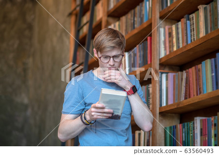 Young man in eyewear reading a book and loooking involved Young man in eyewear reading a book and loooking involved 63560493