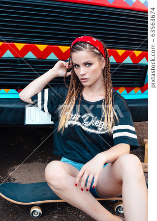 Beautiful smiling girl, with many pigtails wearing in shirt and shorts sitting on skateboard on the street 63563674