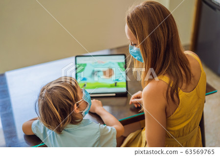 Boy studying online at home using a tablet. Mom helps him learn. Mom and son in medical masks to Boy studying online at home using a tablet. Mom helps him learn. Mom and son in medical masks to 63569765