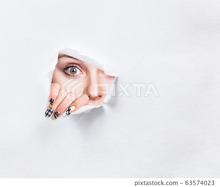 Surprised woman peeps into the slit of white paper. 63574023
