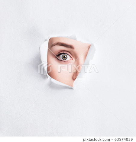 Surprised woman peeps into the slit of white paper. 63574039