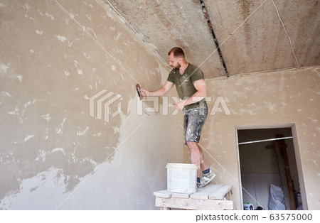 Builder is standing on wooden stand and working with spatula with plaster at the wall in small room 63575000