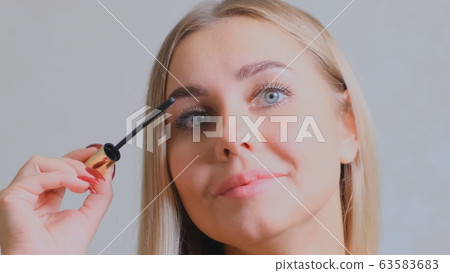 Pretty blond woman applying mascara make-up on her eyelashes in front of mirror. Beauty and makeup Pretty blond woman applying mascara make-up on her eyelashes in front of mirror. Beauty and makeup 63583683
