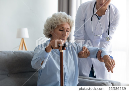 Female nurse help senior lady stand up from couch 63586289