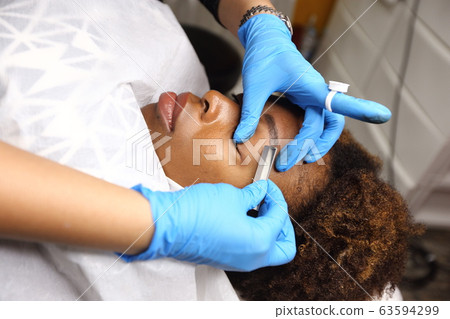 Professional Beautician woman applying Tottoo, Brow Microblading to Black african customer eyebrows to make permanent color makeup or tattooing line look real in beauty salon, copy space for text log Professional Beautician woman applying Tottoo, Brow Microblading to Black african customer eyebrows to make permanent color makeup or tattooing line look real in beauty salon, copy space for text log 63594299