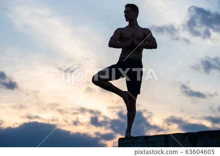 Fit young man practices sun salutation yoga on the edge of cliff at sunset 63604605