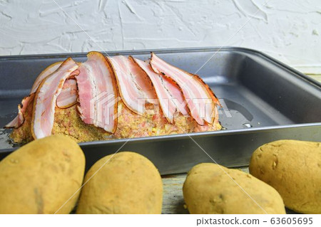 Meat loaf with slices of bacon. Home-made meat loaf on roasting pan. Potatoes foregraund. 63605695