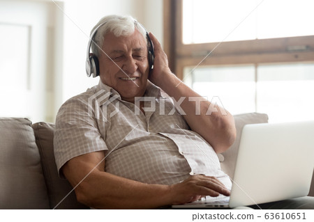 Smiling older man wearing wireless headphones enjoying music, using laptop 63610651