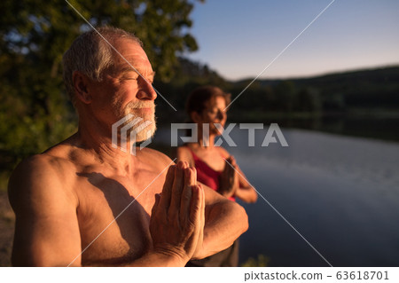 Senior couple in swimsuit standing by lake outdoors at dusk doing yoga. Senior couple in swimsuit standing by lake outdoors at dusk doing yoga. 63618701