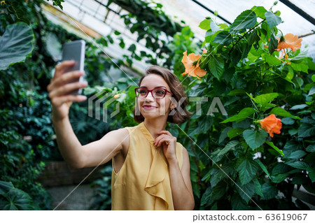 Young woman with smartphone standing in botanical garden, taking selfie. 63619067