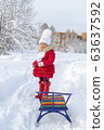 Winter fun. Beautiful little girl in a red coat and mittens smiles and holds a sled on a rope. Snow covered path, trees and snowdrifts. Frosty snowy weather. Clear winter day. Pleasure at fresh air 63637592
