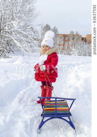 Winter fun. Beautiful little girl in a red coat and mittens smiles and holds a sled on a rope. Snow covered path, trees and snowdrifts. Frosty snowy weather. Clear winter day. Pleasure at fresh air 63637592
