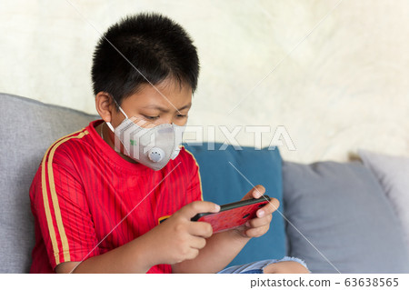 Asian boy with protective mask playing game on cell phone at home. Asian boy with protective mask playing game on cell phone at home. 63638565