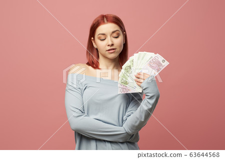 Ginger woman holding euro banknotes in hands while standing in studio Ginger woman holding euro banknotes in hands while standing in studio 63644568