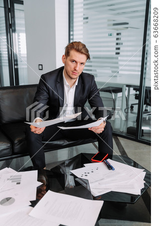 a young stylish businessman is sitting on a sofa in a modern office and holding important documents in his hands a young stylish businessman is sitting on a sofa in a modern office and holding important documents in his hands 63668209