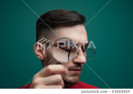Portrait of a young handsome guy looking through a magnifying glass on a green background 63668984