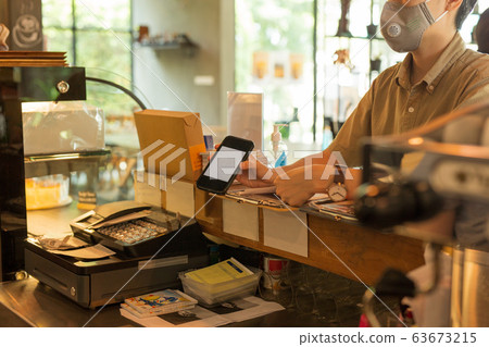 Woman customer with protective mask paying bill by cell phone in cafe. 63673215