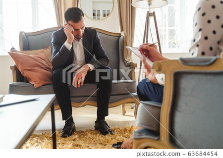 Low-spirited dark-haired man sitting on a sofa Low-spirited dark-haired man sitting on a sofa 63684454