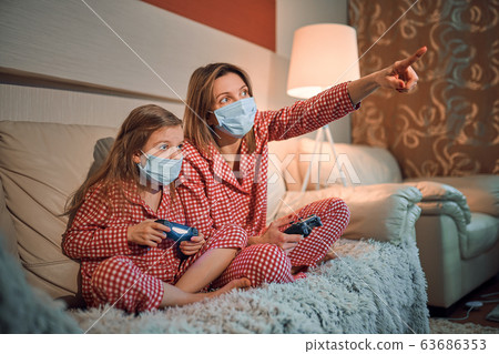 Woman and young girl wearing pajamas and medical protective masks sitting on sofa in living room with video game controllers at home isolation auto quarantine, covid-19 Woman and young girl wearing pajamas and medical protective masks sitting on sofa in living room with video game controllers at home isolation auto quarantine, covid-19 63686353