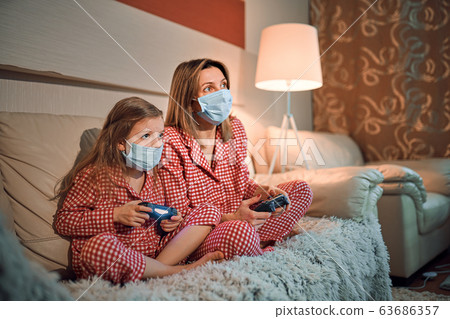 Woman and young girl wearing pajamas and medical protective masks sitting on sofa in living room with video game controllers at home isolation auto quarantine, covid-19 Woman and young girl wearing pajamas and medical protective masks sitting on sofa in living room with video game controllers at home isolation auto quarantine, covid-19 63686357