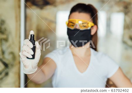 A girl in a protective mask, glasses and gloves spray an antiseptic to prevent infection. 63702402