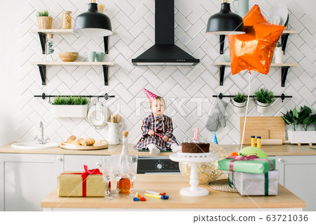 childhood, people and celebration concept - happy baby girl in pink birthday hat sitting in the kitchen on birthday party at home. Table with birthday cake, presents, air balloons and party horns childhood, people and celebration concept - happy baby girl in pink birthday hat sitting in the kitchen on birthday party at home. Table with birthday cake, presents, air balloons and party horns 63721036