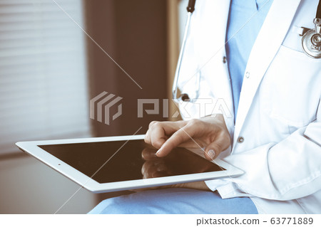 Woman doctor using white tablet computer while sitting at chair in hospital, close-up 63771889