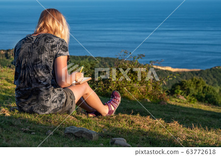 A young, blonde girl writes her thoughts in a personal diary on the mountain with the view to the sea. Enjoy your time and moments. Travel and relaxation A young, blonde girl writes her thoughts in a personal diary on the mountain with the view to the sea. Enjoy your time and moments. Travel and relaxation 63772618